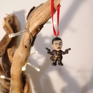 May include: A brown plastic figurine of a person holding two guns, hanging from a red ribbon on a branch. The figurine is a Christmas ornament.