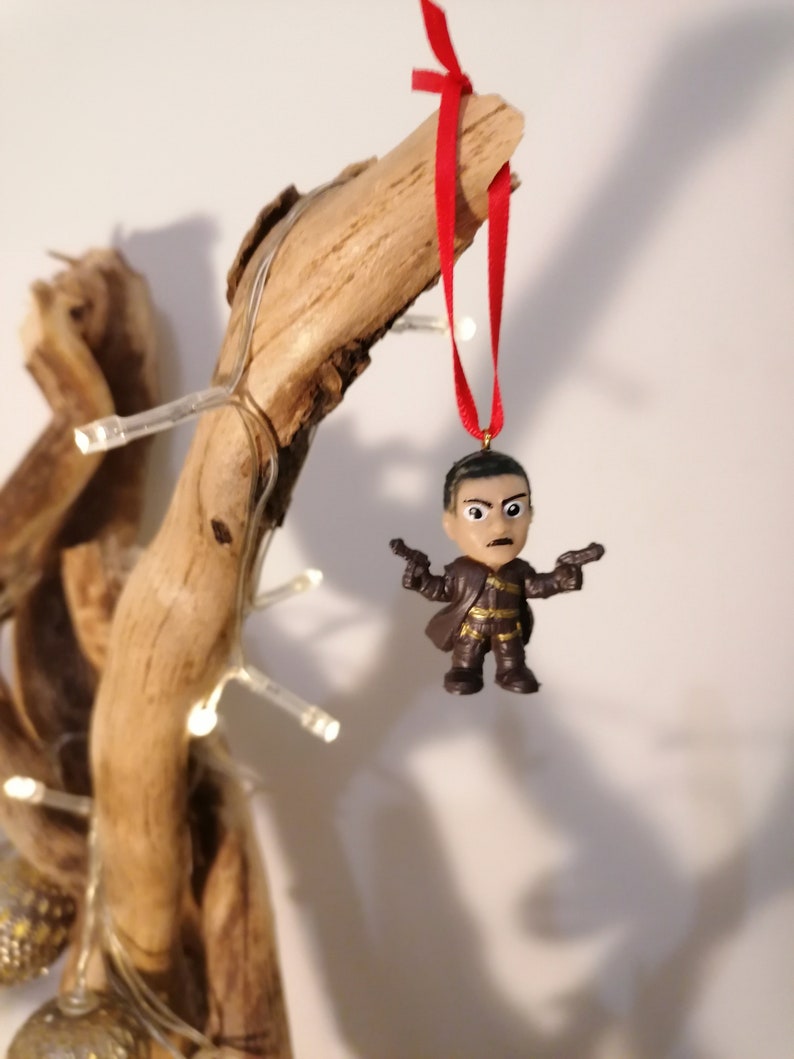 May include: A brown plastic figurine of a person holding two guns, hanging from a red ribbon on a branch. The figurine is a Christmas ornament.