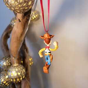 May include: A red string hangs from a branch with a small plastic figurine of Woody from Toy Story. Woody is wearing his signature cowboy hat, vest, and jeans. He is holding his arm out and his legs are bent as if he is walking.