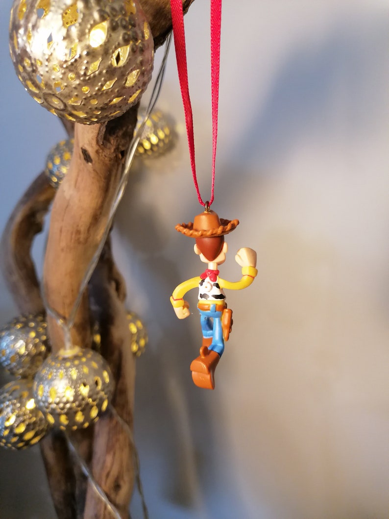 May include: A red string hangs from a branch with a small plastic figurine of Woody from Toy Story. Woody is wearing his signature cowboy hat, vest, and jeans. He is holding his arm out and his legs are bent as if he is walking.