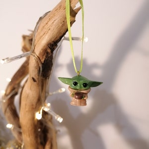 May include: A green and brown Yoda ornament with a green ribbon hanging from a branch. The ornament is holding a small brown object.