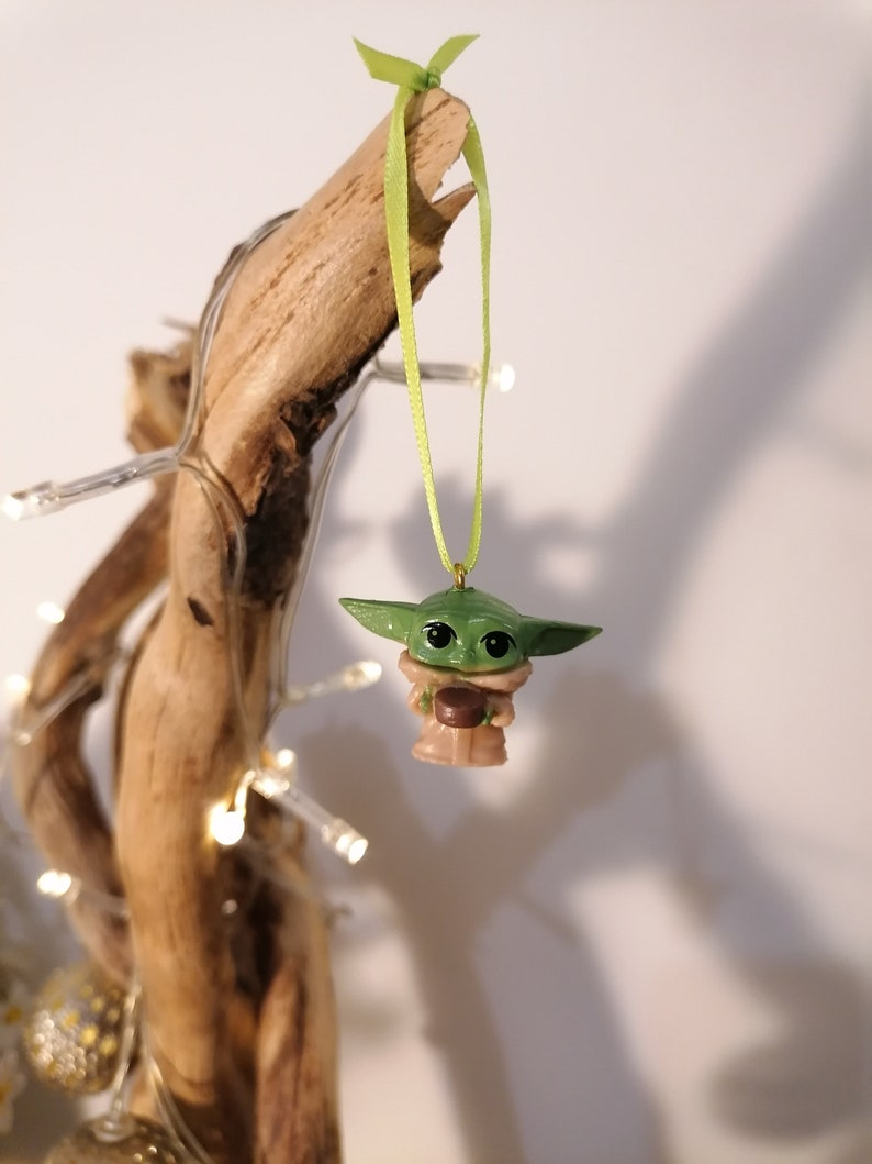 May include: A green and brown Yoda ornament with a green ribbon hanging from a branch. The ornament is holding a small brown object.