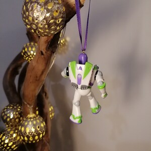 May include: A white and green Buzz Lightyear toy ornament hanging from a purple ribbon on a branch with gold and white decorative balls.