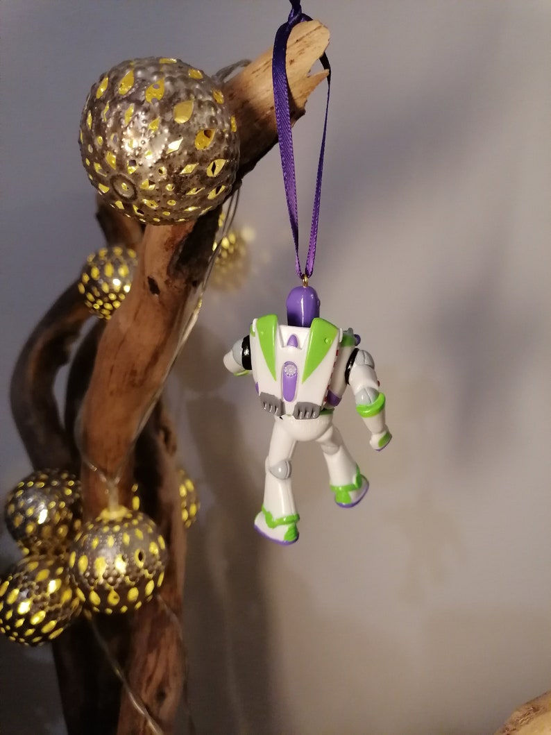 May include: A white and green Buzz Lightyear toy ornament hanging from a purple ribbon on a branch with gold and white decorative balls.