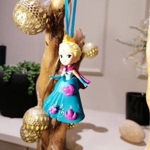 K&ouml;nnte beinhalten: Eine blaue und violette Plastikfigur einer Prinzessin mit blonden Haaren, die an einem blauen Band an einem Holzast h&auml;ngt. Die Prinzessin tr&auml;gt ein blaues Kleid mit violetten Blumen und eine Krone.