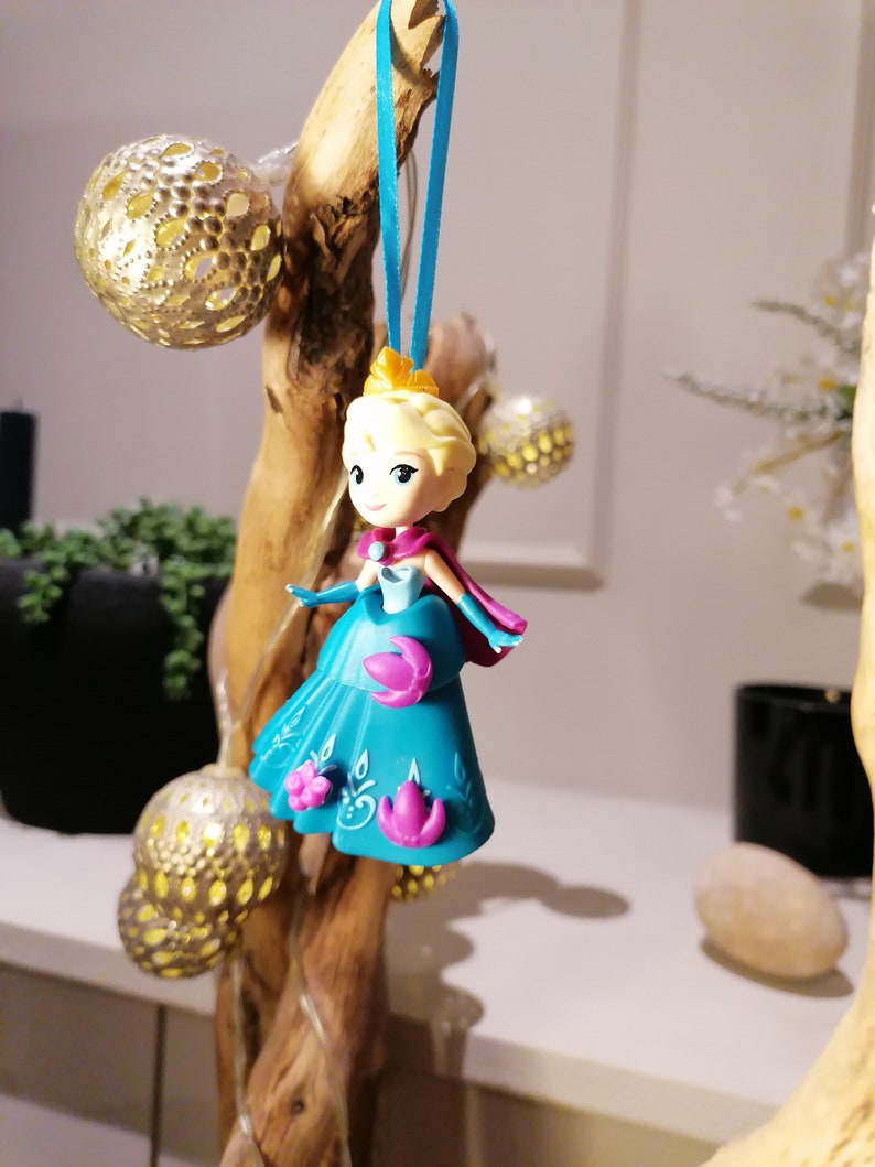 K&ouml;nnte beinhalten: Eine blaue und violette Plastikfigur einer Prinzessin mit blonden Haaren, die an einem blauen Band an einem Holzast h&auml;ngt. Die Prinzessin tr&auml;gt ein blaues Kleid mit violetten Blumen und eine Krone.