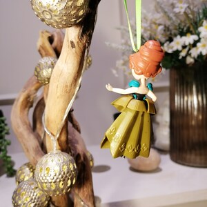 May include: A gold and green plastic figurine of a princess with red hair hanging from a green ribbon on a branch with silver and gold ornaments.