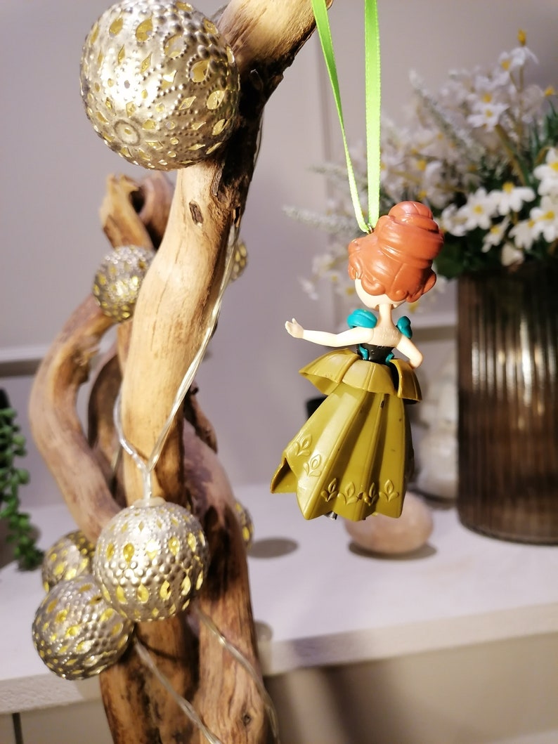May include: A gold and green plastic figurine of a princess with red hair hanging from a green ribbon on a branch with silver and gold ornaments.