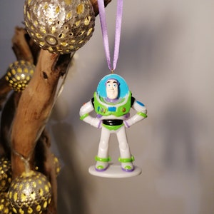 Puede incluir: Un adorno navideño de plástico con Buzz Lightyear de la franquicia Toy Story. El adorno es blanco, verde y morado con una visera azul. Buzz Lightyear está de pie con las manos en las caderas.