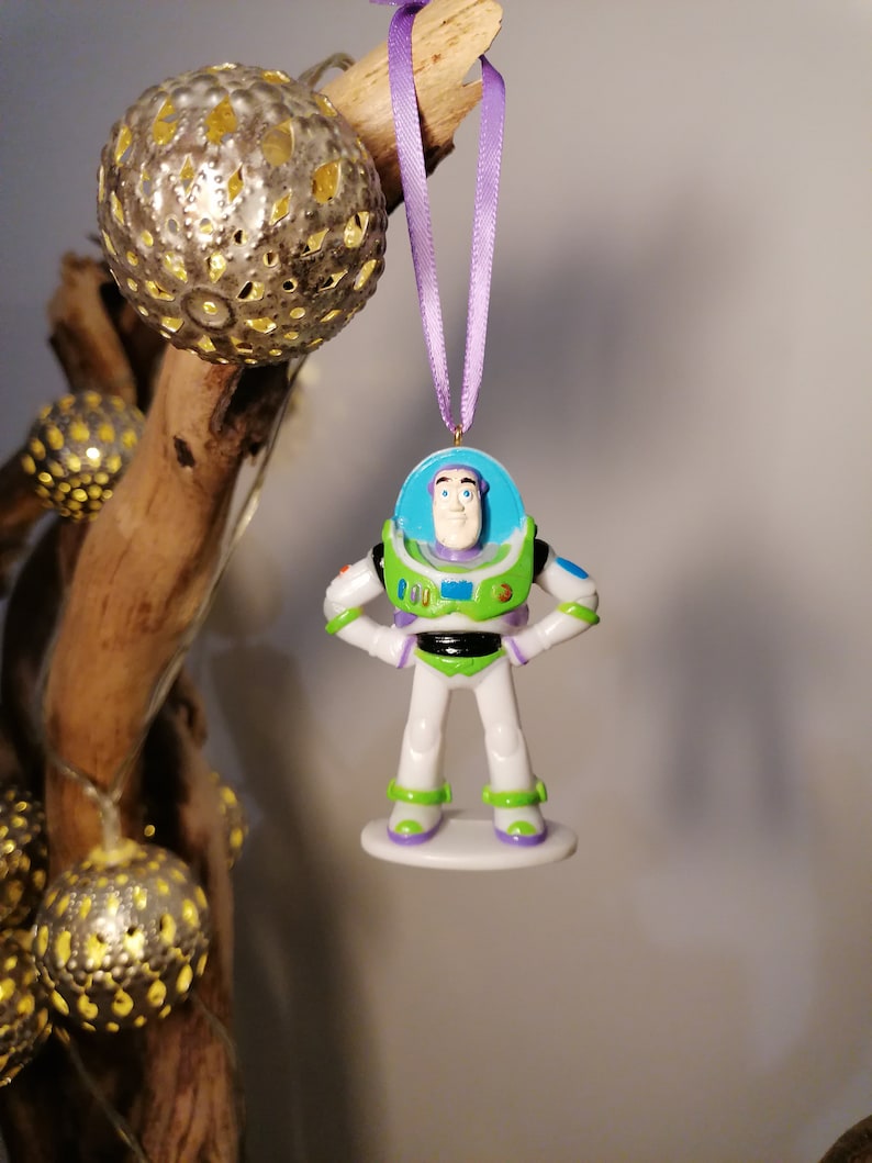 Puede incluir: Un adorno navide&ntilde;o de pl&aacute;stico con Buzz Lightyear de la franquicia Toy Story. El adorno es blanco, verde y morado con una visera azul. Buzz Lightyear est&aacute; de pie con las manos en las caderas.