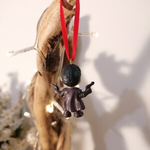 May include: A small, black and gray figurine of a person holding two guns, hanging from a red ribbon. The figurine is a Christmas ornament.