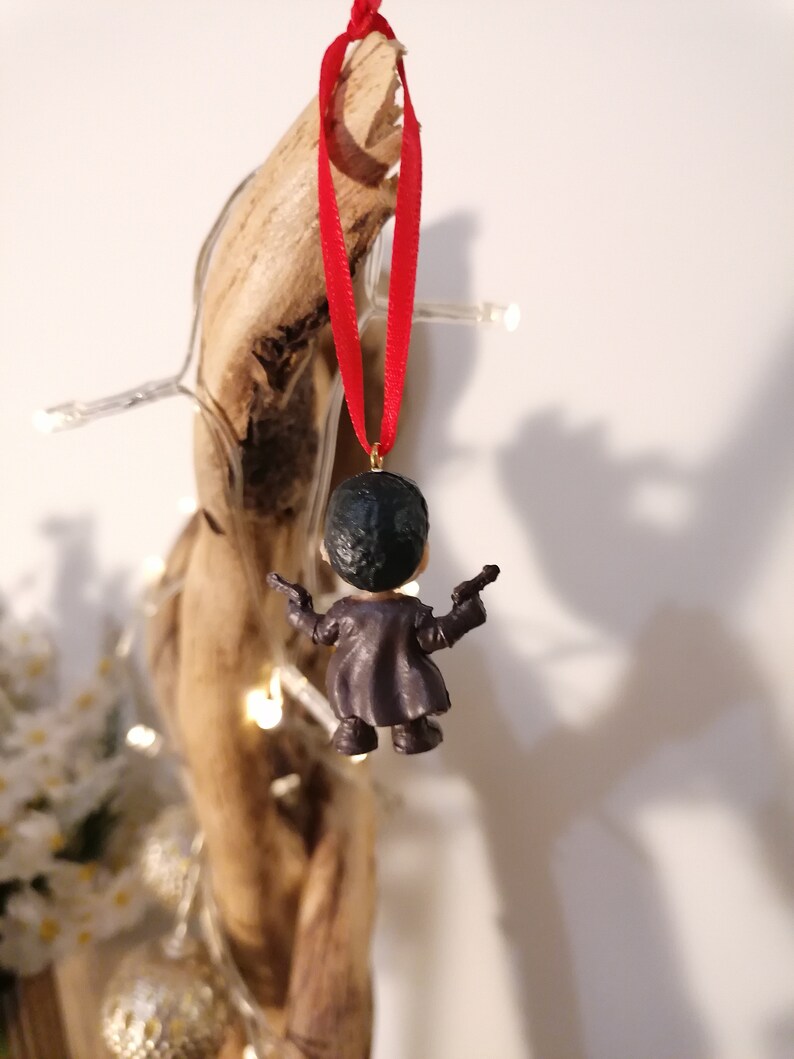May include: A small, black and gray figurine of a person holding two guns, hanging from a red ribbon. The figurine is a Christmas ornament.