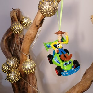 May include: A green and blue toy car with a figure of Woody from Toy Story driving it. The car is hanging from a branch with a string of lights. The lights are silver and have a yellow glow.