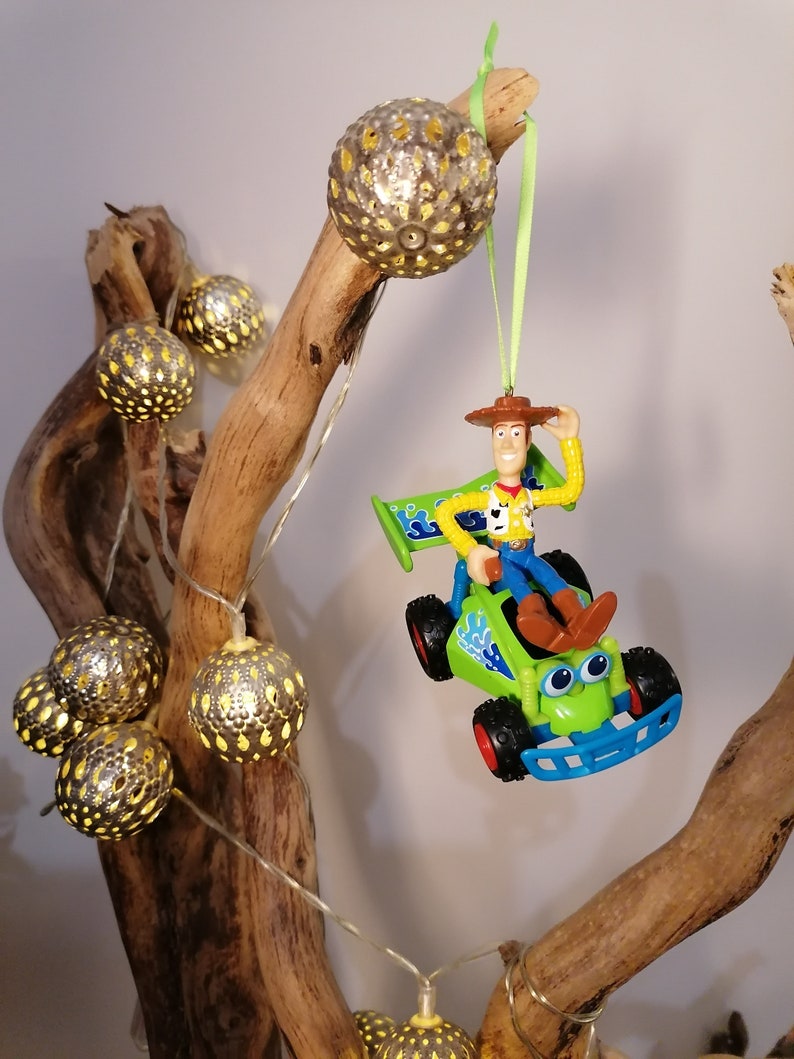 May include: A green and blue toy car with a figure of Woody from Toy Story driving it. The car is hanging from a branch with a string of lights. The lights are silver and have a yellow glow.