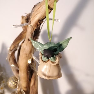May include: A green and brown Yoda ornament hanging from a branch with a green ribbon. The ornament is made of polymer clay and is about 3 inches tall.
