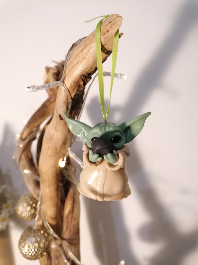 May include: A green and brown Yoda ornament hanging from a branch with a green ribbon. The ornament is made of polymer clay and is about 3 inches tall.