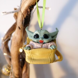 May include: A green and gray character ornament with large eyes and a pink mouth, sitting in a yellow bag, hanging from a green ribbon.
