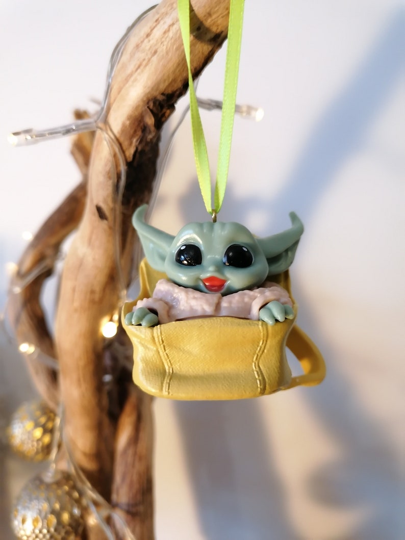 May include: A green and gray character ornament with large eyes and a pink mouth, sitting in a yellow bag, hanging from a green ribbon.