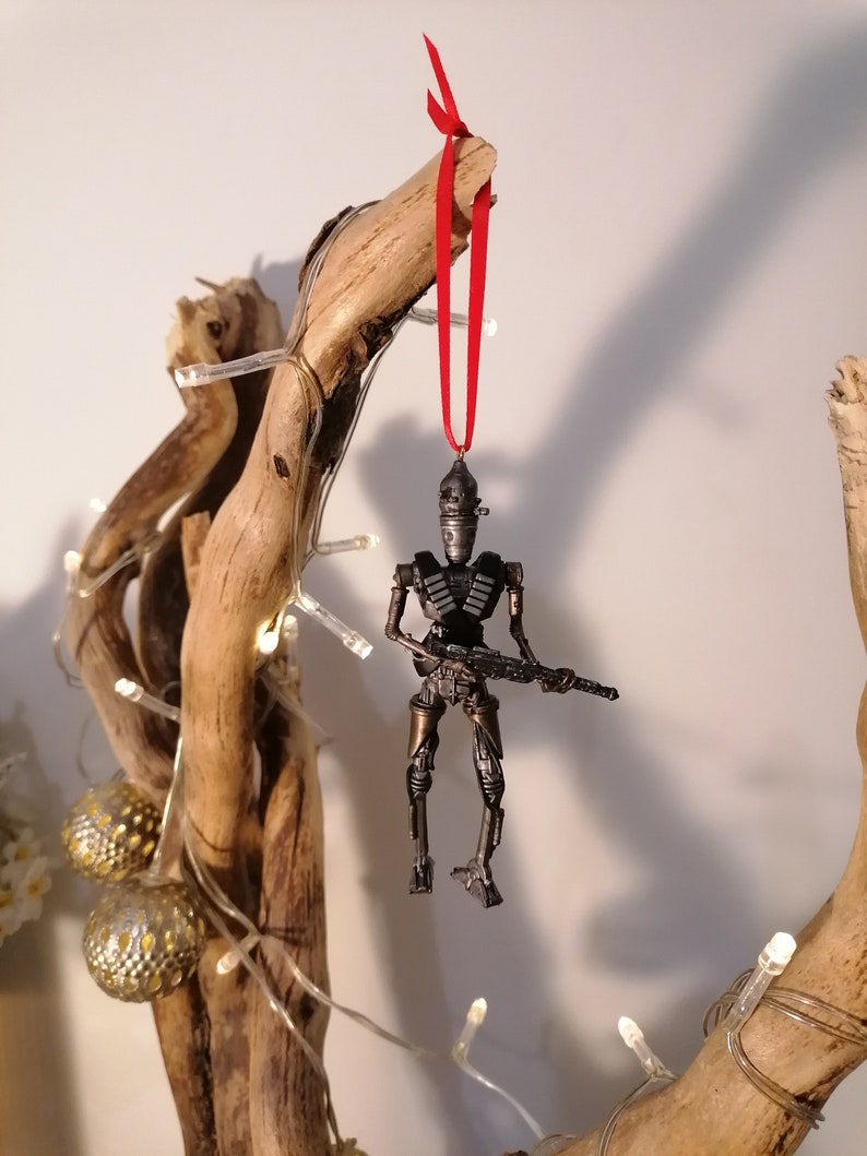 Puede incluir: Un adorno de robot gris met&aacute;lico con una pistola colgando de una cinta roja en una rama con luces blancas.