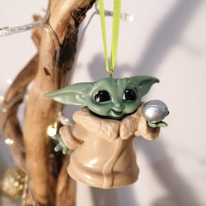 May include: A green and brown Christmas ornament featuring a character from the Star Wars franchise, holding a silver ball.