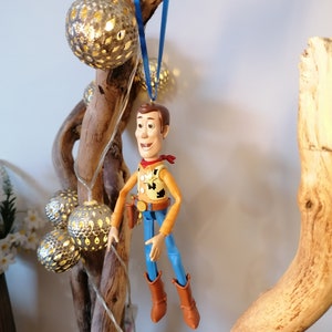 Puede incluir: Una figura de juguete de pl&aacute;stico de Woody de Toy Story colgada de una cinta azul en una rama de madera a la deriva con luces de cadena y bolas de metal decorativas.