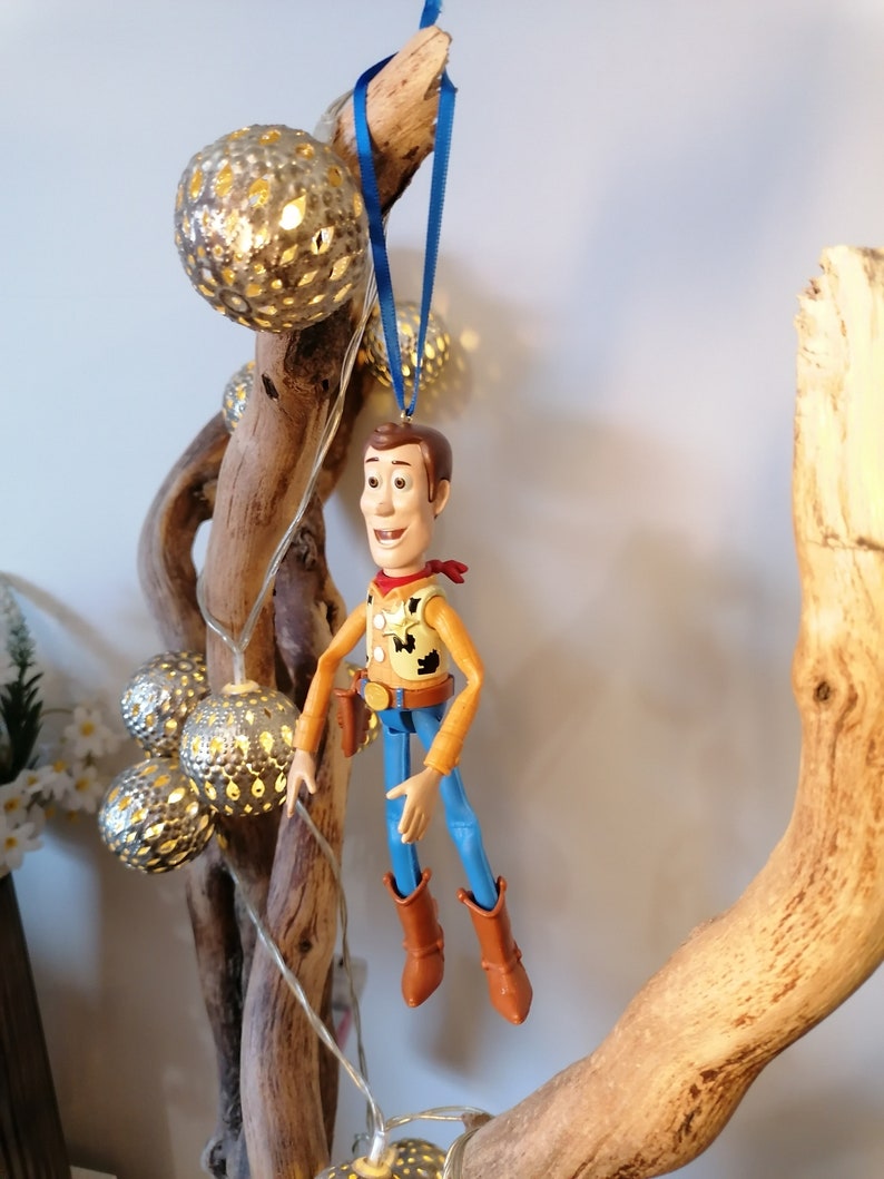 Puede incluir: Una figura de juguete de pl&aacute;stico de Woody de Toy Story colgada de una cinta azul en una rama de madera a la deriva con luces de cadena y bolas de metal decorativas.