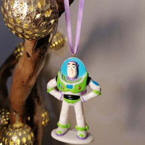 Puede incluir: Un adorno de pl&aacute;stico de Buzz Lightyear de la franquicia Toy Story. El adorno es blanco con detalles verdes y azules. Buzz Lightyear lleva su uniforme de ranger espacial caracter&iacute;stico y tiene una cinta morada atada a la parte superior de su cabeza.