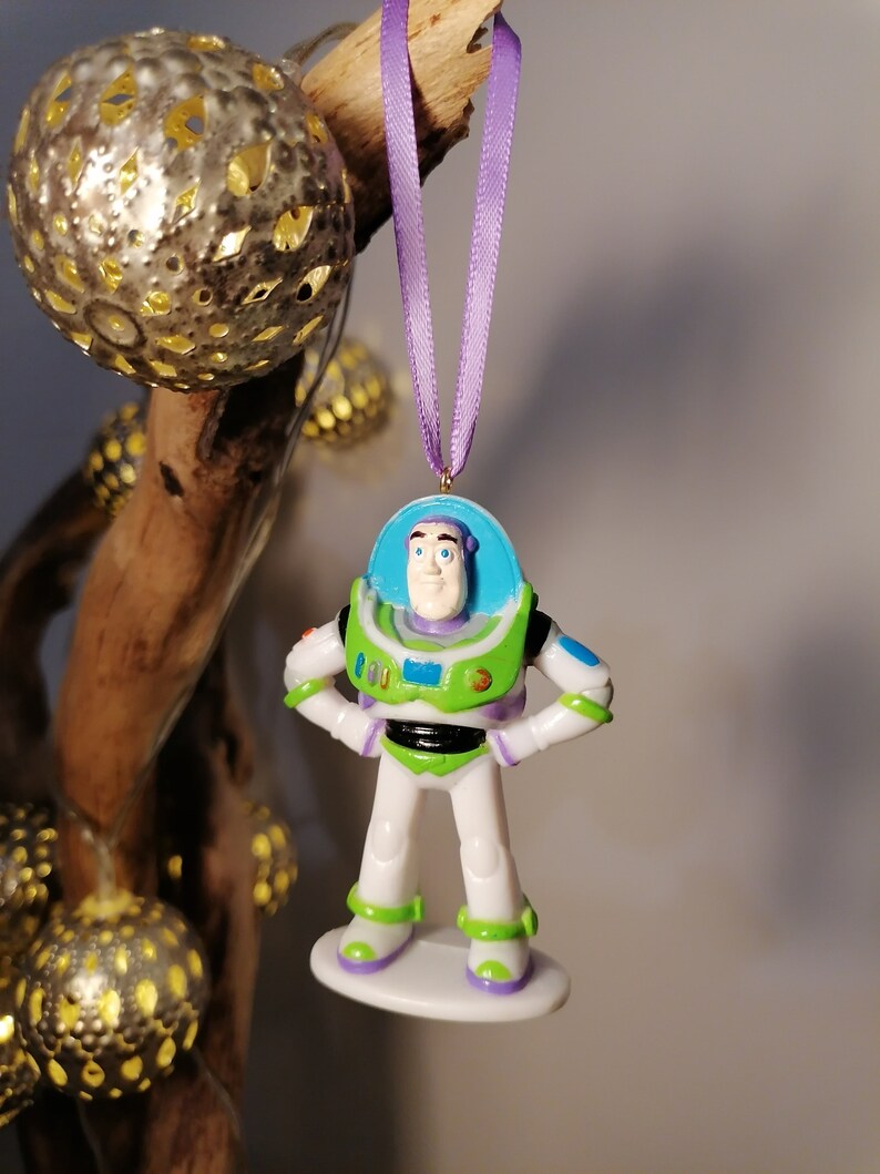 Puede incluir: Un adorno de pl&aacute;stico de Buzz Lightyear de la franquicia Toy Story. El adorno es blanco con detalles verdes y azules. Buzz Lightyear lleva su uniforme de ranger espacial caracter&iacute;stico y tiene una cinta morada atada a la parte superior de su cabeza.