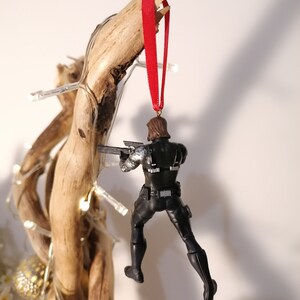 May include: A black and silver action figure of a superhero hanging from a red ribbon. The figure is holding a gun and is posed in a dynamic action pose.