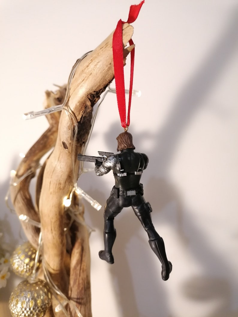 May include: A black and silver action figure of a superhero hanging from a red ribbon. The figure is holding a gun and is posed in a dynamic action pose.