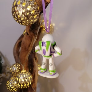 Puede incluir: Un adorno navide&ntilde;o de Buzz Lightyear blanco y verde con una cinta morada colgando de una rama. El adorno est&aacute; hecho de pl&aacute;stico y tiene un dise&ntilde;o detallado.