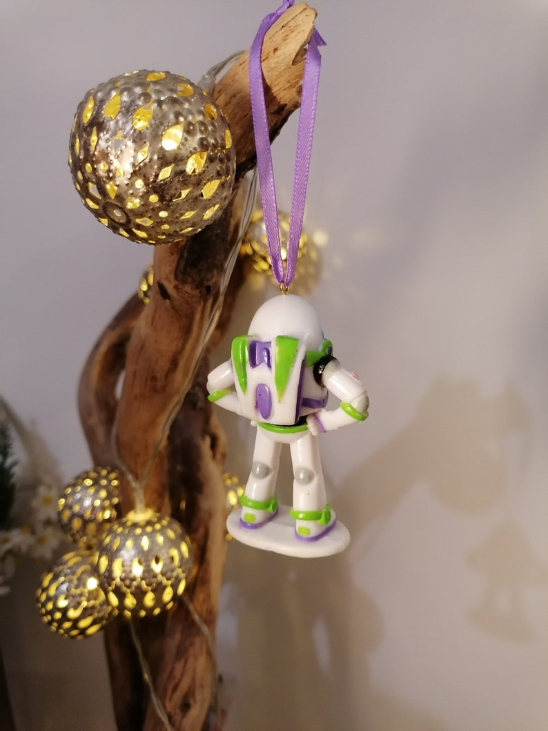 Puede incluir: Un adorno navide&ntilde;o de Buzz Lightyear blanco y verde con una cinta morada colgando de una rama. El adorno est&aacute; hecho de pl&aacute;stico y tiene un dise&ntilde;o detallado.