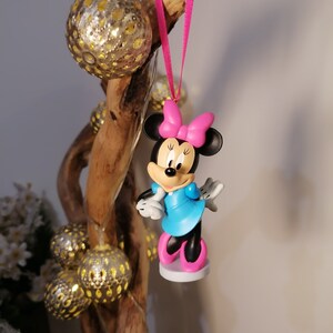 May include: A pink and blue Minnie Mouse Christmas ornament with a pink bow. The ornament is hanging from a pink ribbon on a wooden branch.