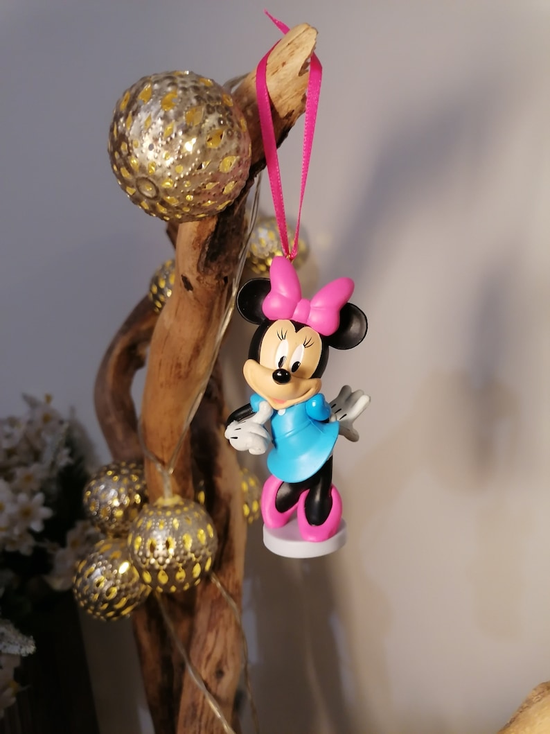 May include: A pink and blue Minnie Mouse Christmas ornament with a pink bow. The ornament is hanging from a pink ribbon on a wooden branch.