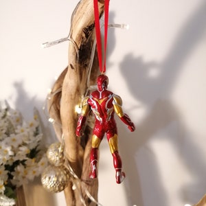 May include: Red and gold Iron Man Christmas ornament hanging from a branch with a red ribbon. The ornament is a detailed figurine of Iron Man in his red and gold suit.