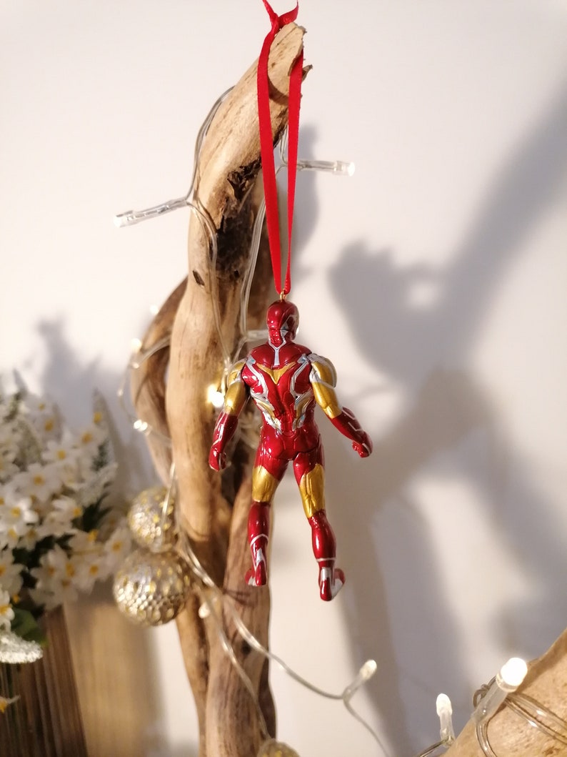 May include: Red and gold Iron Man Christmas ornament hanging from a branch with a red ribbon. The ornament is a detailed figurine of Iron Man in his red and gold suit.