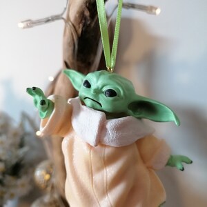 May include: A green and beige Christmas ornament featuring a character from the Star Wars franchise. The character is wearing a beige robe with a white collar and has large ears.