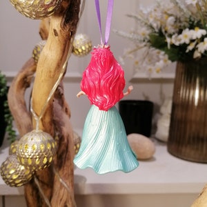 May include: A teal blue and pink plastic figurine of a mermaid hanging from a purple ribbon. The mermaid has long red hair and is wearing a teal blue dress.