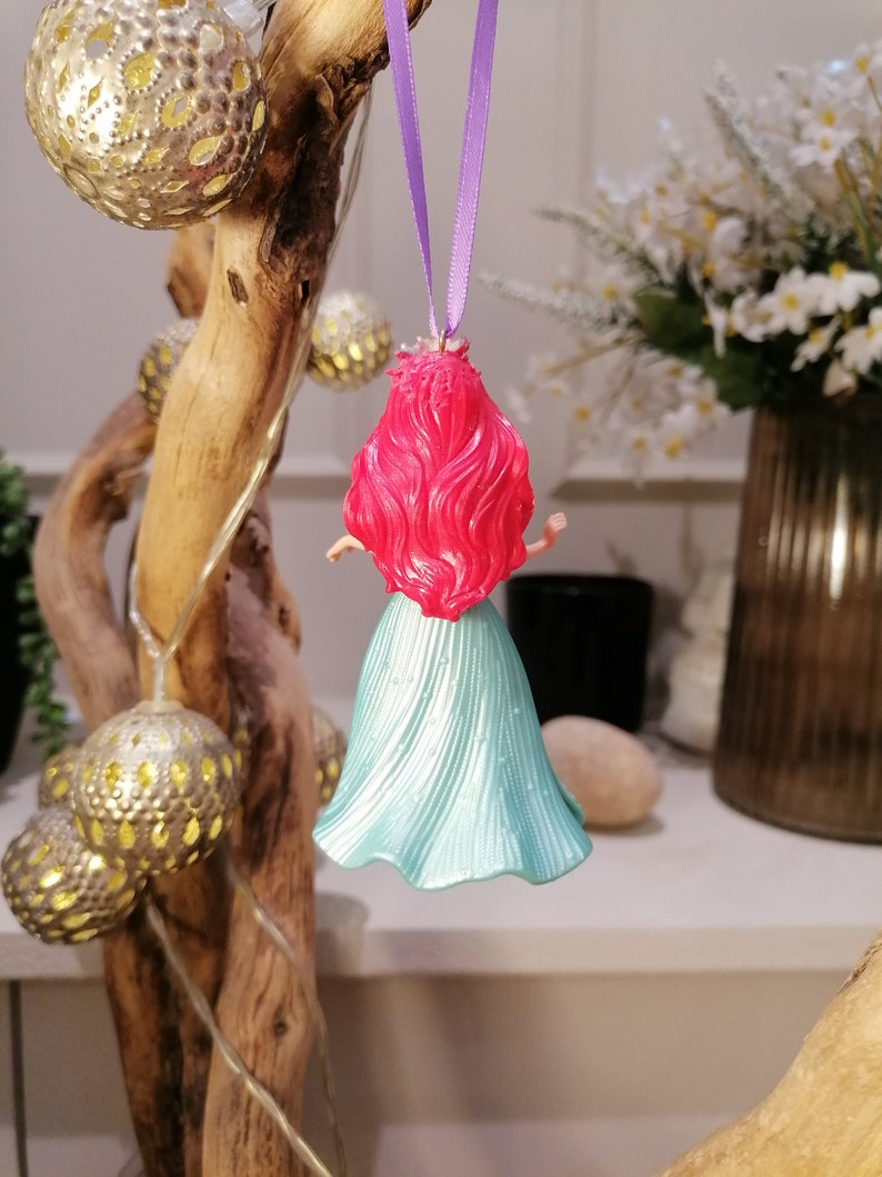 May include: A teal blue and pink plastic figurine of a mermaid hanging from a purple ribbon. The mermaid has long red hair and is wearing a teal blue dress.