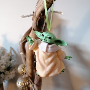 May include: A green and beige Christmas ornament featuring a character from the Star Wars franchise. The character is wearing a beige robe and has large ears. The ornament is hanging from a green ribbon.