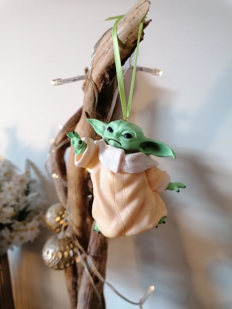 May include: A green and beige Christmas ornament featuring a character from the Star Wars franchise. The character is wearing a beige robe and has large ears. The ornament is hanging from a green ribbon.