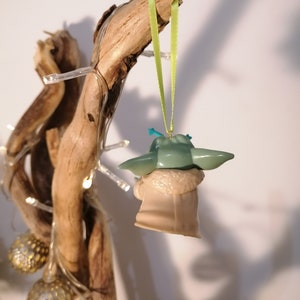 May include: A green and beige Baby Yoda Christmas ornament hanging from a branch with a green ribbon. The ornament is made of plastic and has a detailed design.
