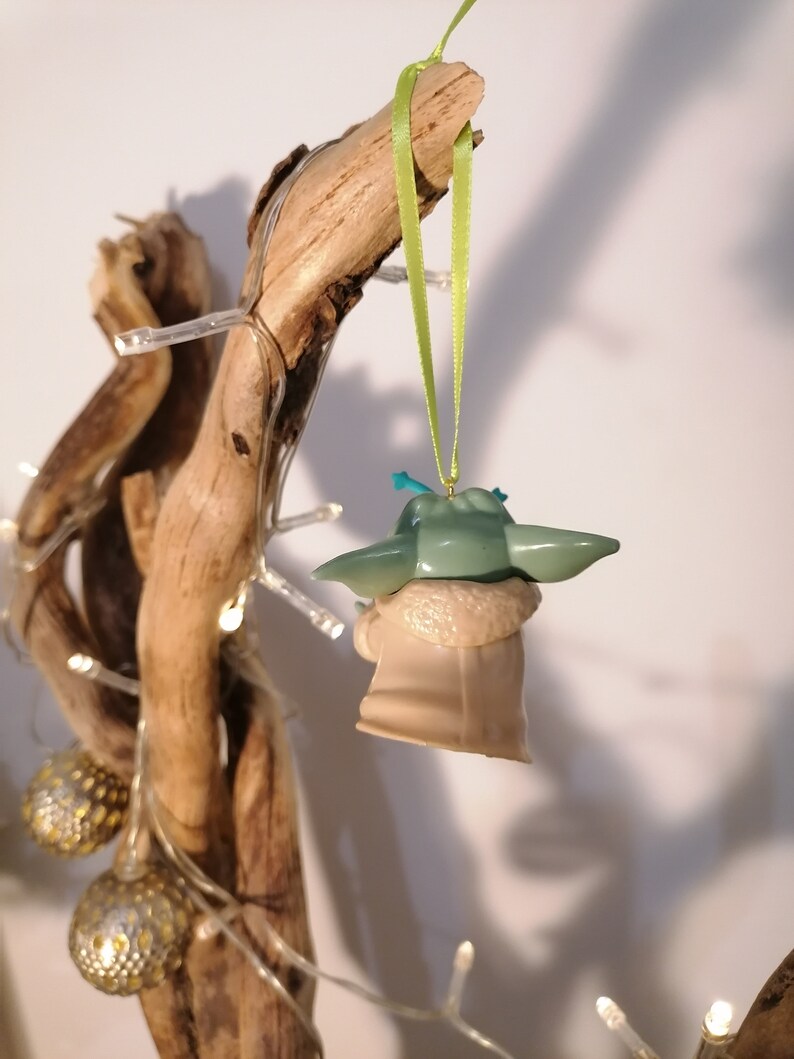 May include: A green and beige Baby Yoda Christmas ornament hanging from a branch with a green ribbon. The ornament is made of plastic and has a detailed design.