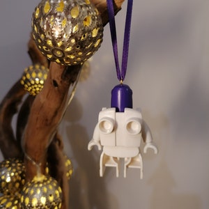May include: White Lego minifigure ornament with a purple head and a purple ribbon hanging from a branch with string lights.