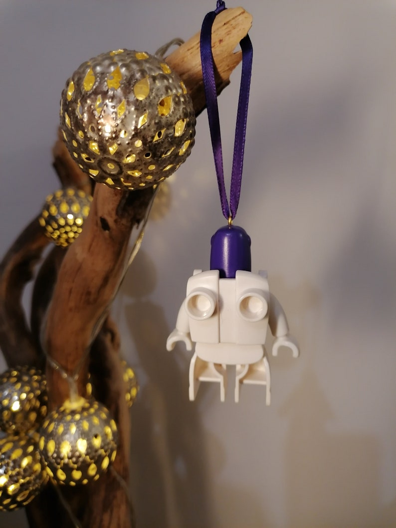 May include: White Lego minifigure ornament with a purple head and a purple ribbon hanging from a branch with string lights.