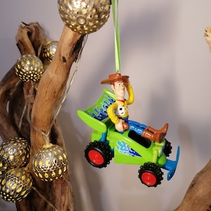 May include: A green toy car with a figure of Woody from Toy Story driving. The car has blue flames on the side and red wheels. The ornament is hanging from a green ribbon.