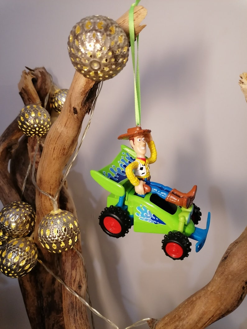 May include: A green toy car with a figure of Woody from Toy Story driving. The car has blue flames on the side and red wheels. The ornament is hanging from a green ribbon.