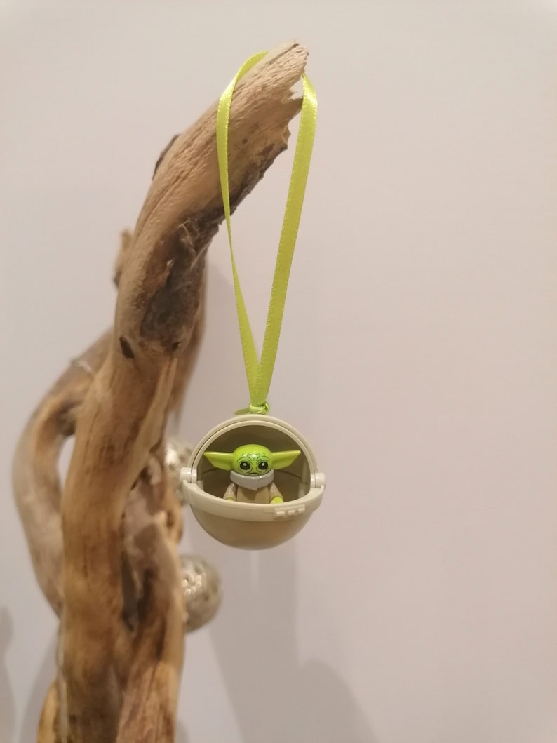 May include: A green and tan Baby Yoda ornament in a pod, hanging from a light green ribbon. The ornament is suspended from a piece of driftwood against a white background. The Baby Yoda figure is wearing a mask.