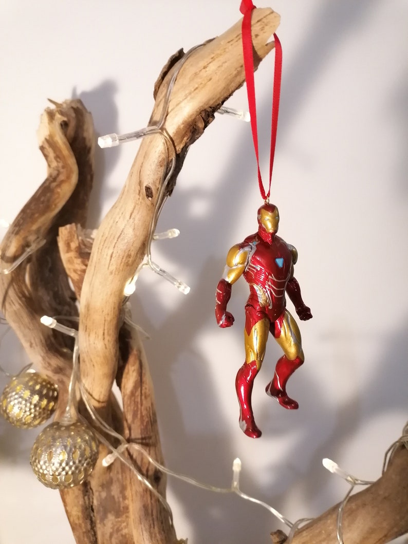 May include: Red and gold Iron Man Christmas ornament hanging from a branch with white lights.