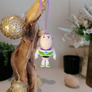 Puede incluir: Adorno navideño de Buzz Lightyear morado y blanco con una cinta morada. El adorno está colgado de una rama.